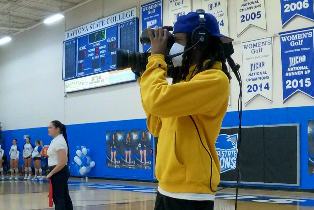 student in yellow sweater filming a volleyball game