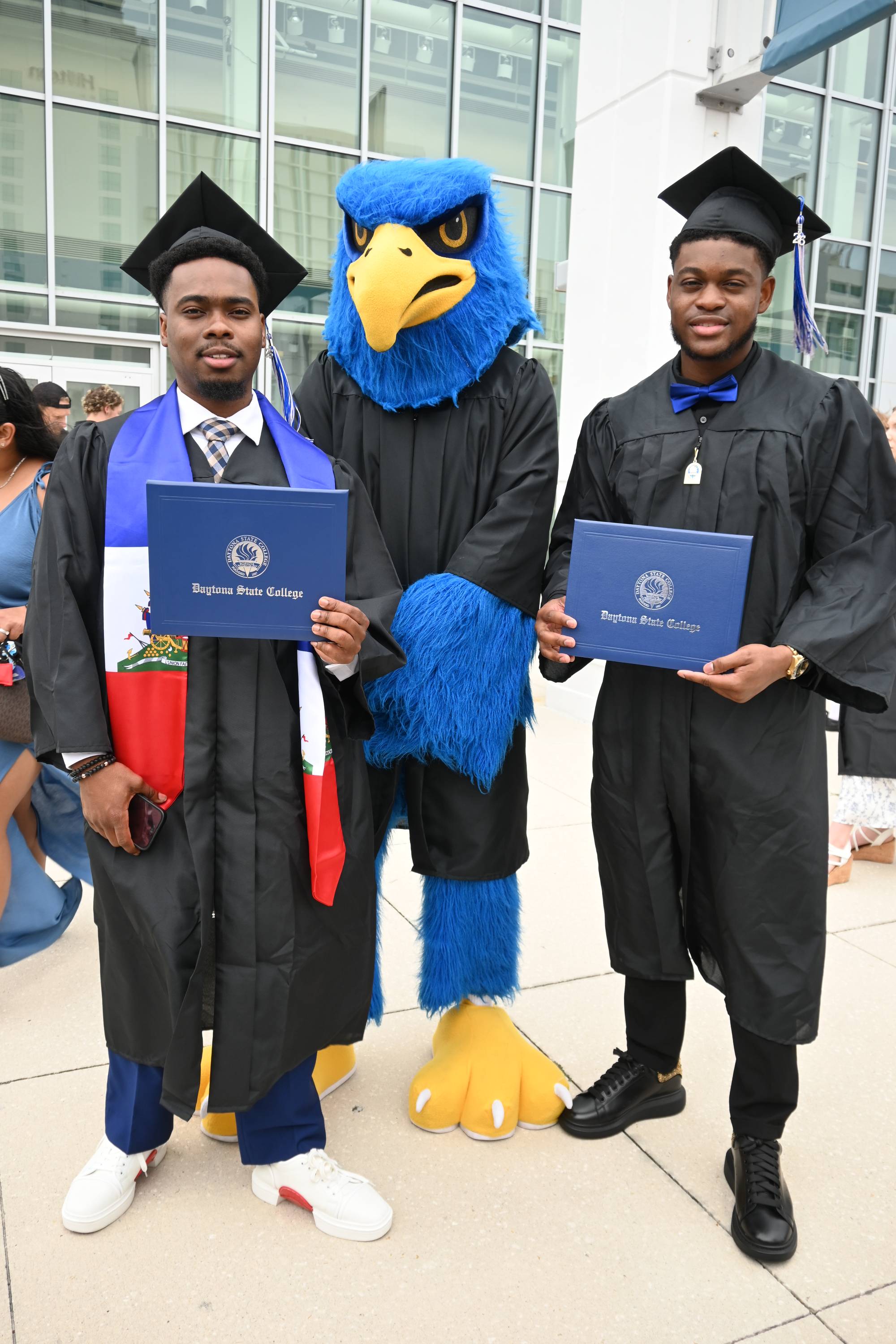 Two graduates posing with Freddie the Falcon at Commencement. 